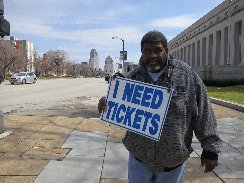 Photo of a ticket scalper holding a sign that reads "I Need Tickets".